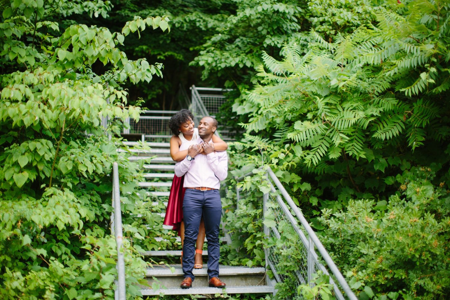A couple standing on a metal staircase with green trees surrounding them at Evergreen Brickworks. The woman has her arms around her fiancés shoulders.