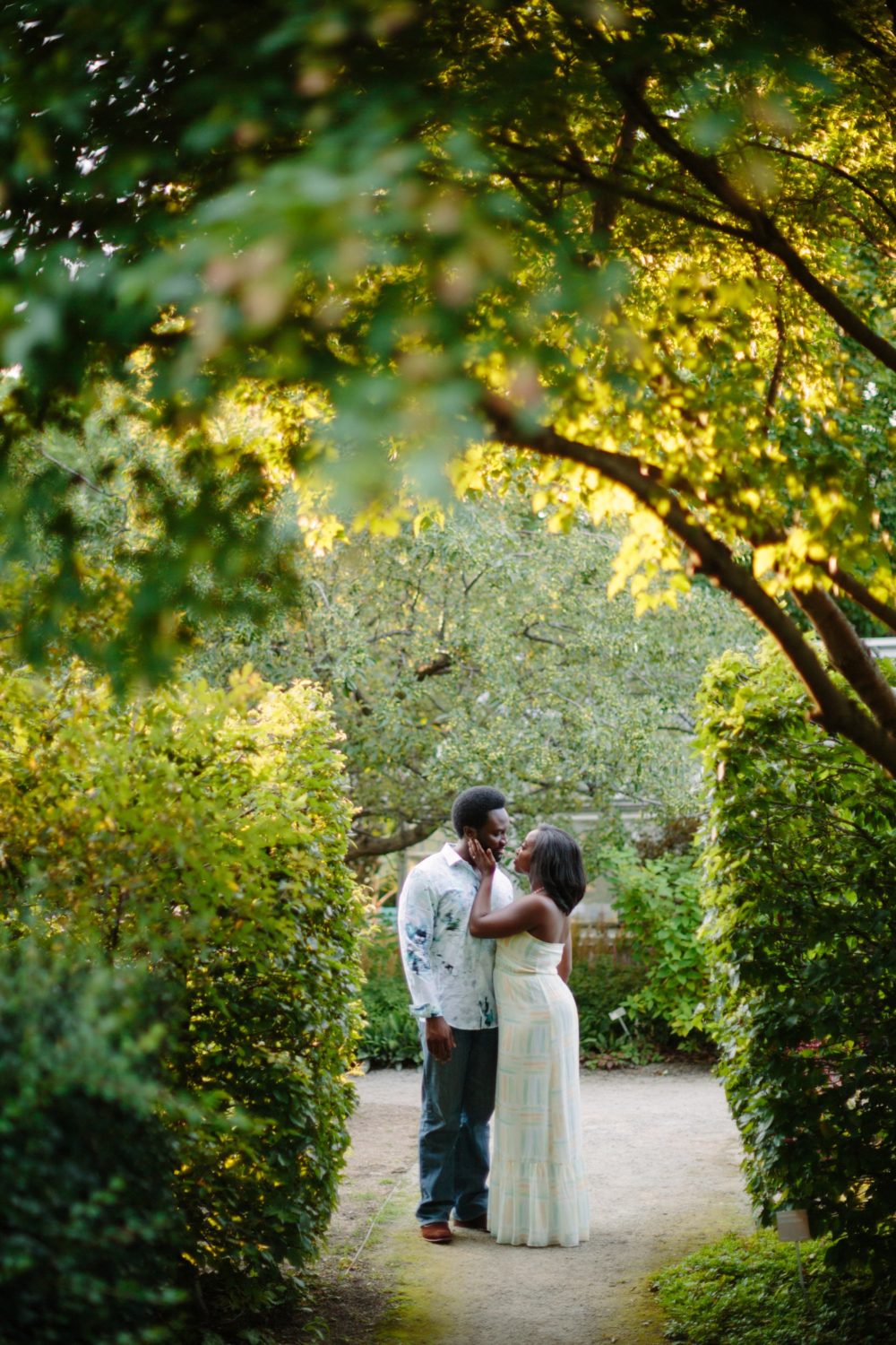 A couple standing facing each other in the Toronto Botanical Gardens. The woman is holding her fiancés cheek with er hand. Trees and shrubs frame the couple.