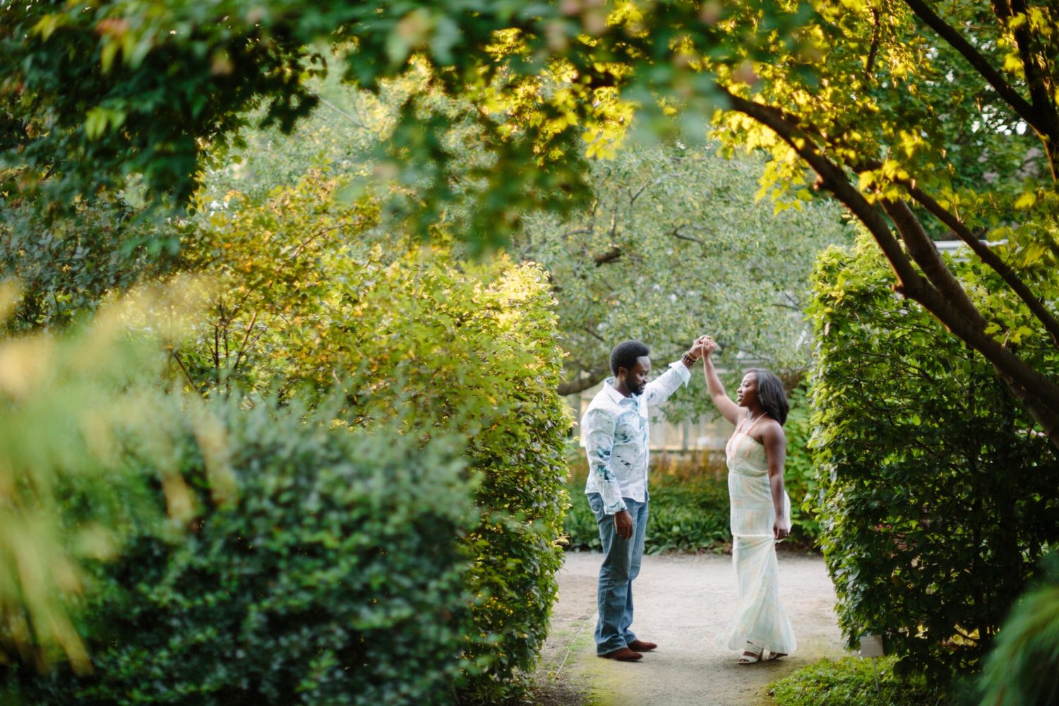 A couple holding hands above their heads facing each other. Green shrubs and trees frame the couple.